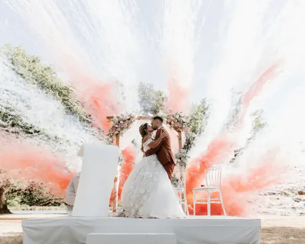 Un couple s'embrasse sous une arche fleurie, entouré de fumée rose et blanche. Ces photos mariage capturent un moment magique.