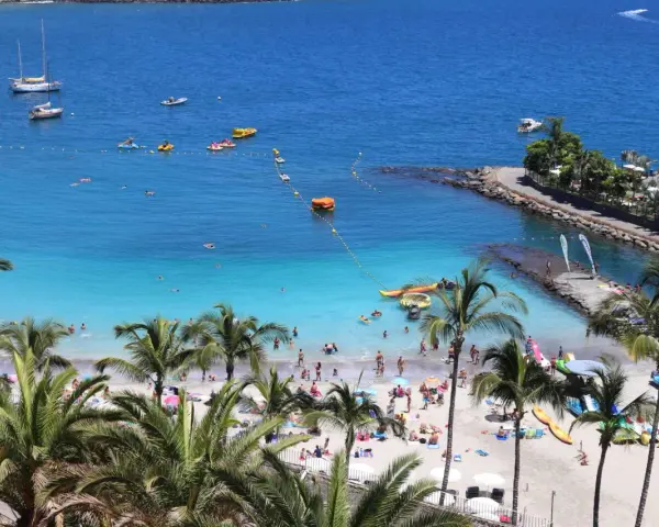 Słoneczne gran canaria plaże z palmami, turkusową wodą i ludźmi odpoczywającymi na piasku. Widać łodzie i sprzęt wodny.