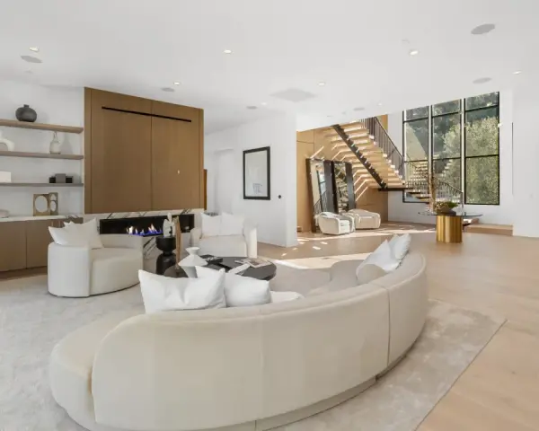 Soggiorno luminoso con camino moderno, divano curvo e poltrone bianche. Scale a vista e grandi finestre definiscono l'arredamento minimal chic.