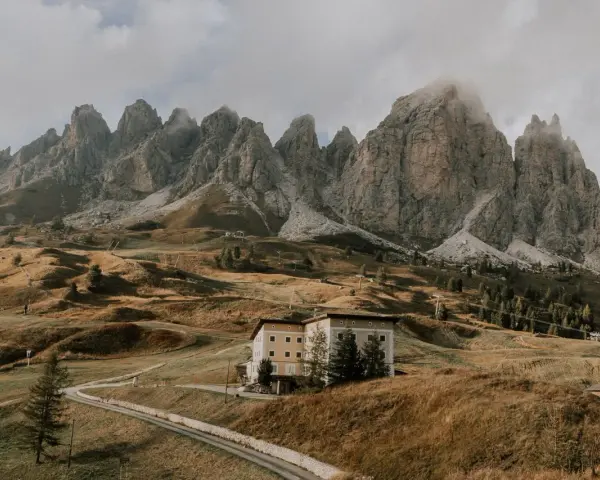 Malownicze **Val di Funes** z górami w tle, hotel na wzgórzu i kręta droga.