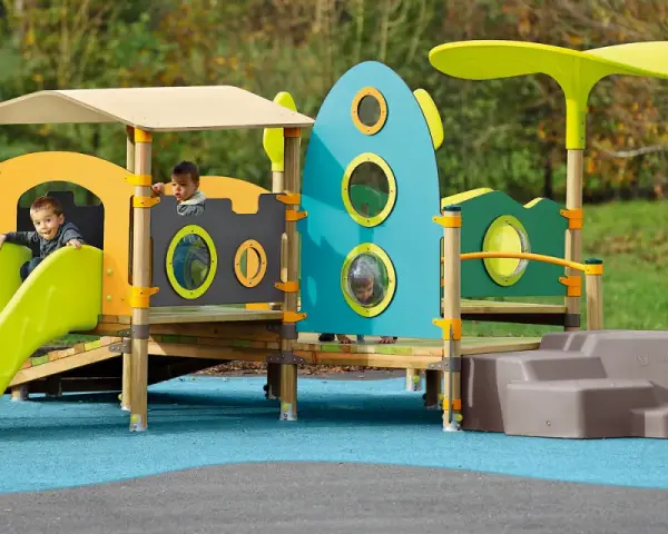 Des enfants jouent sur une aire de jeux colorée, respectant la réglementation aire de jeux extérieur. Un toboggan vert vif et une structure inspirée d'une fusée bleue.