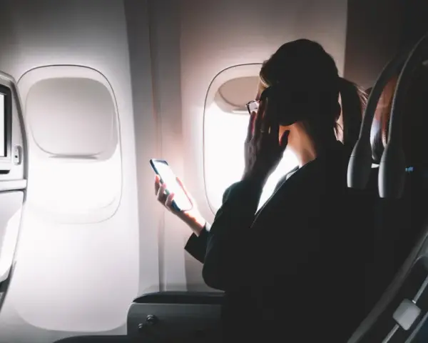 Kobieta w okularach trzyma telefon, zastanawiając się, czy w samolocie jest wifi. Obok ekran rozrywki.