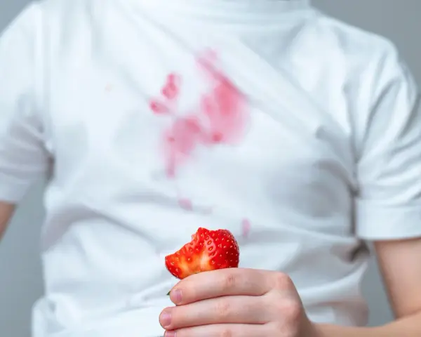 Dziecko trzyma kawałek truskawki, a na białej koszulce widać czerwone plamy z truskawek.