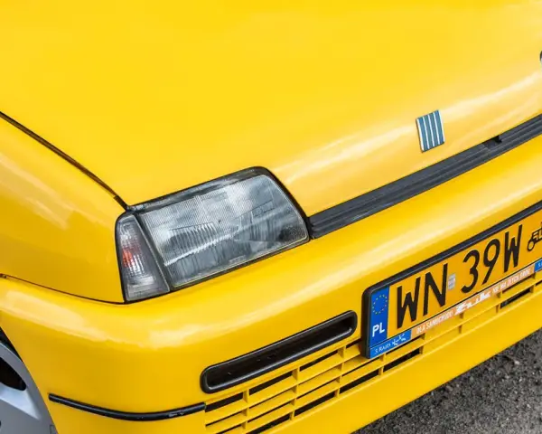 Żółte tablice rejestracyjne na błyszczącym, żółtym samochodzie Fiat. Widoczny fragment maski, reflektor i zderzak.