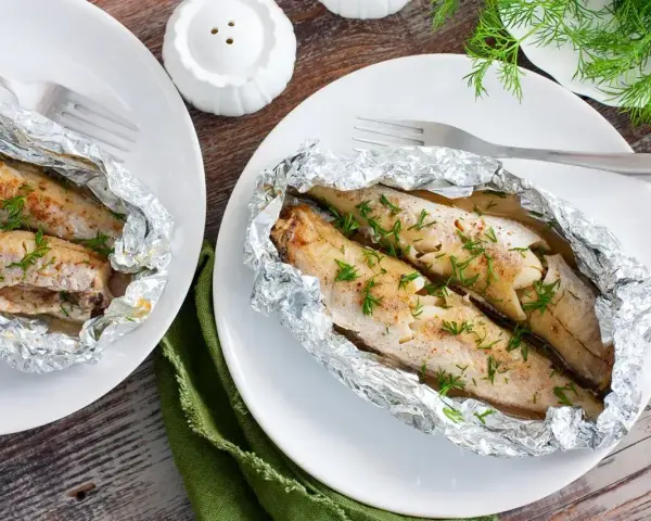 Dwa porcje soczystego mintaja z piekarnika, zapieczonego w folii aluminiowej, posypane świeżym koperkiem.