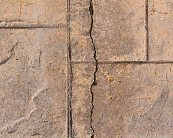 A prominent crack runs through a stamped concrete slab foundation, revealing sand and smaller fissures.