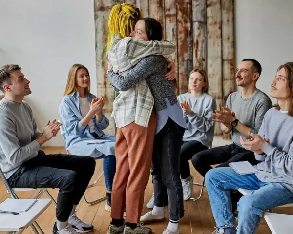 Grupa ludzi siedzi w kręgu, klaszcząc. Dwie osoby w centrum mocno się przytulają. Co to aa? Wsparcie i akceptacja.