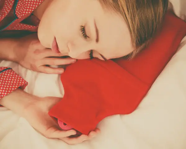 Une femme dort paisiblement, blottie contre une bouillotte rouge. Une astuce pour savoir comment faire une bouillotte avec une serviette pour se réchauffer.