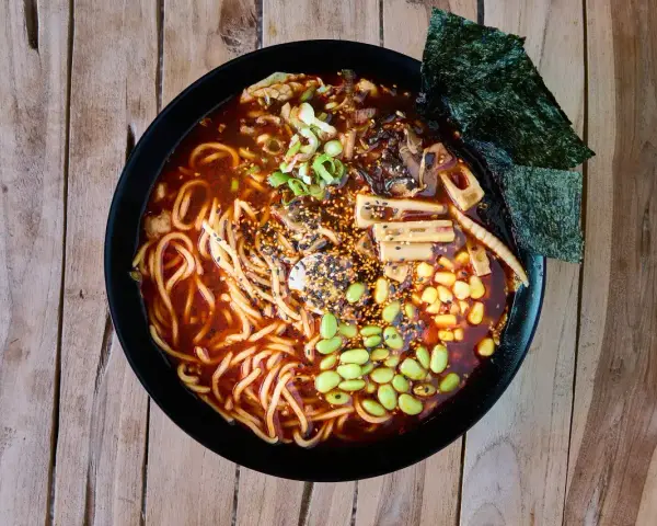 Miseczka aromatycznych dań z makaronem ramen, pełna edamame, kukurydzy, grzybów i nori, na drewnianym tle.