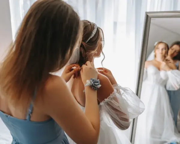 Świadek na weselu pomaga pannie młodej poprawić welon i biżuterię. W lustrze widać odbicie pary młodej.