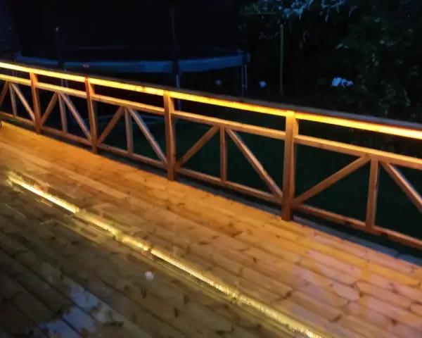 Träaltan med staket upplyst av varma decklights. En grill står på altanen.