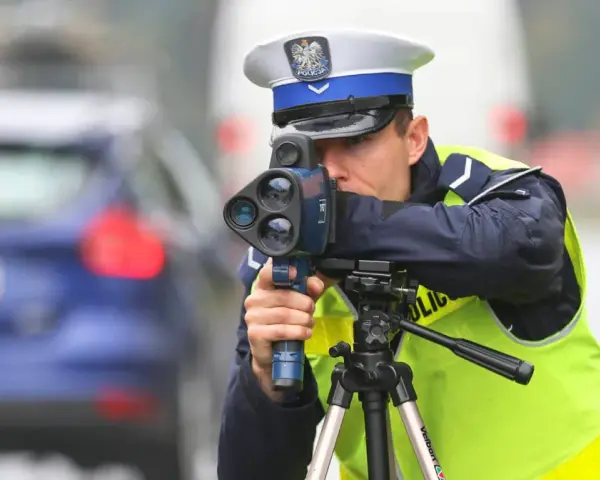 Policjant w kamizelce odblaskowej mierzy prędkość samochodu za pomocą radaru. Trwa dozór policyjny na drodze.