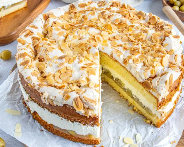 Ein köstlicher Stachelbeer Baiser Kuchen mit Biskuitböden, Sahne und Mandelsplittern. Ein Stück ist bereits herausgeschnitten.