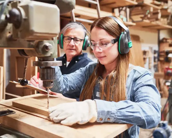 Junge Frau lernt an einer Bohrmaschine, während ein älterer Mann zusieht. Eine gute Ausbildung, welche Ausbildungen dauern 2 Jahre, ist hier vielleicht der Anfang.