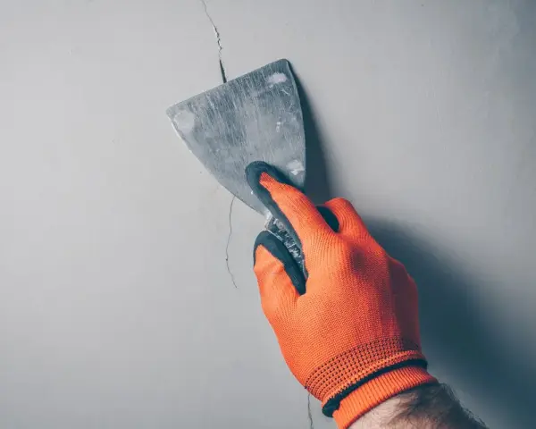 Ręka w pomarańczowej rękawicy zacierająca pęknięcie w ścianie szpachlą. Idealne rozwiązanie, czym wypełnić głębokie pęknięcia w ścianie.