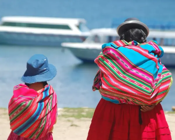 Zwei cholitas escaladoras, gekleidet in bunte Ponchos und Hüte, blicken auf Boote auf einem See.
