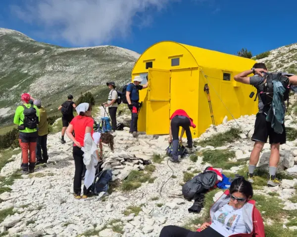Gruppo di escursionisti vicino a un giallo blockhaus Majella, su un sentiero roccioso con montagne verdi sullo sfondo.