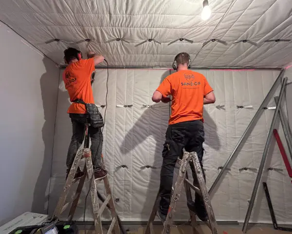 Dwóch mężczyzn w pomarańczowych koszulkach montuje materiał izolacyjny, pokazując, jak wygłuszyć ścianę.