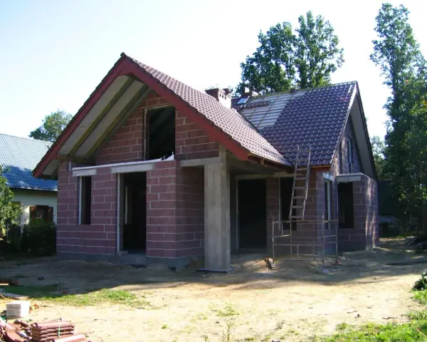 Dom w stanie surowym otwartym, z czerwonej cegły, z częściowo położonym dachem. Wokół plac budowy.