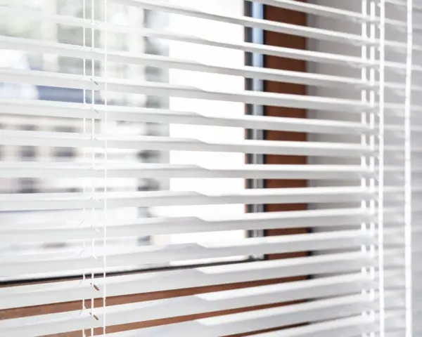 Close-up of white horizontal blinds with visible cords, showing the slats and a hint of the window frame.
