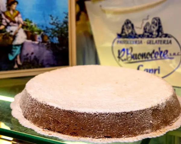 A Torta Caprese dusted with powdered sugar sits on a glass counter, with a painting and shop logo in the background.