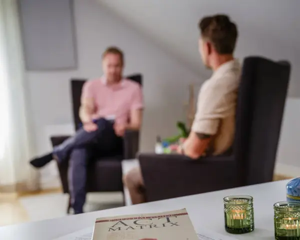 Två män i samtal i en lugn miljö. En bok om acceptance and commitment therapy svenska ligger på bordet.