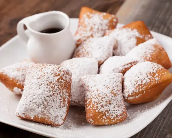 Une assiette de beignets saupoudrés de sucre glace, parfaits pour une recette pâte à beignets salés. Un petit pichet de sauce est à côté.