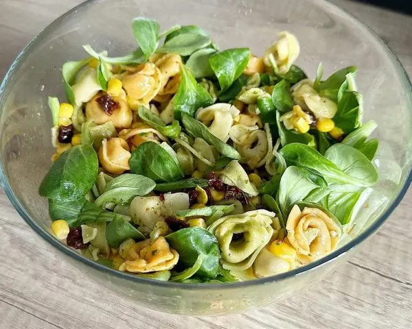 Kolorowa sałatka z tortellini, kukurydzą i suszonymi pomidorami w szklanej misce.