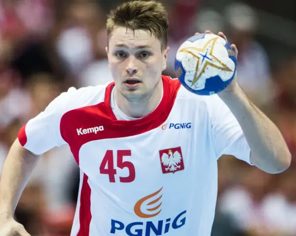 Michał Szyba, numer 45, w białej koszulce z czerwonymi elementami, trzyma piłkę do piłki ręcznej.