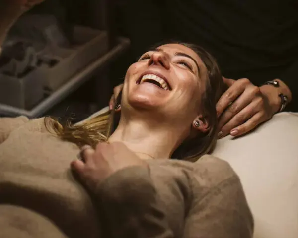 A woman laughs joyfully during a holistic facial treatment, her head tilted back as hands gently support her.