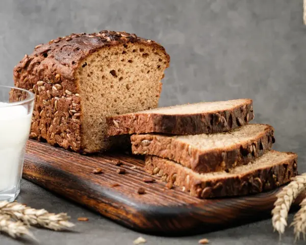 Chleb proteinowy z ziarnami, pokrojony na kromki, obok szklanka mleka i kłosy zboża. Idealny na zdrowe śniadanie.