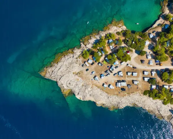 Widok z lotu ptaka na kemping Riviera Makarska. Kamieniste wybrzeże, turkusowa woda i zielone drzewa otaczają liczne przyczepy kempingowe i samochody.