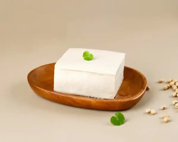Kostka białego tofu na drewnianej miseczce, ozdobiona listkiem. Obok garść ziaren soi. To właśnie co to tofu – produkt z soi.
