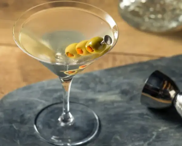 Elegancki martini z wódką, ozdobiony trzema oliwkami na wykałaczce, gotowy do degustacji. Obok metalowy jigger.
