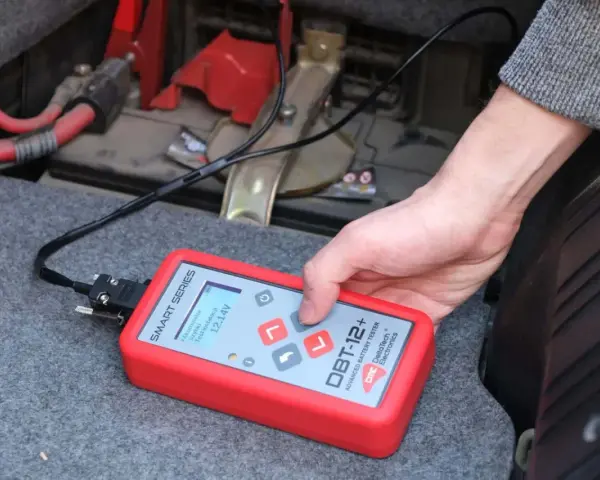 Ręka trzyma tester akumulatora DBT-12+, pokazujący 12.14V. To prosty sposób, jak sprawdzić akumulator w samochodzie.