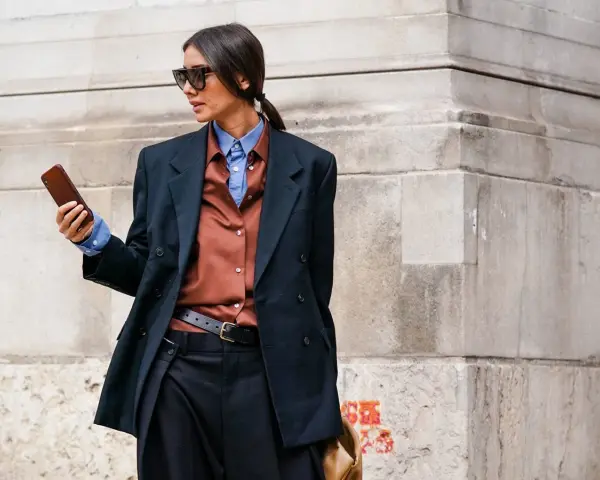 Mujer con estilo combinando traje negro con camisa marrón y azul, gafas de sol y móvil en mano.