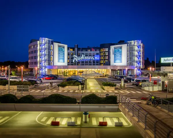 Nowoczesny hotel Radisson Blu Hamburg Airport nocą, z podświetlanym logo i parkingiem pełnym samochodów.