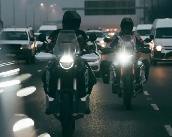 Dwóch motocyklistów w kaskach jedzie w deszczu. Ich motocykle mają włączone światła, a droga polna jest mokra i błyszcząca.