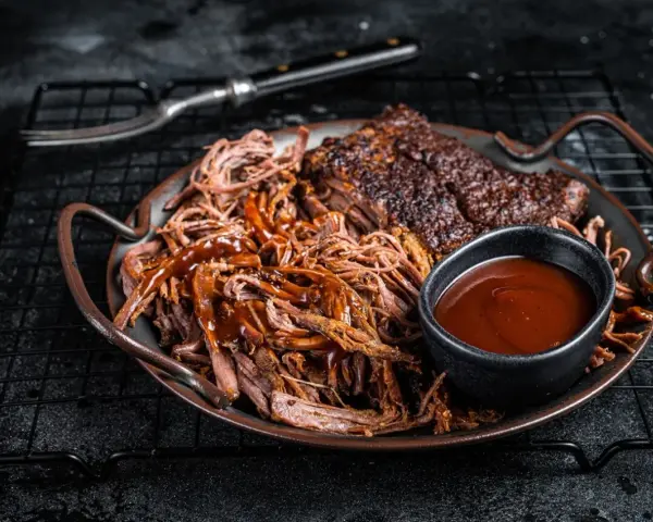 Saftiges pulled pork mit BBQ-Sauce und Beilagen auf einem Teller. Ein Gabel liegt daneben.