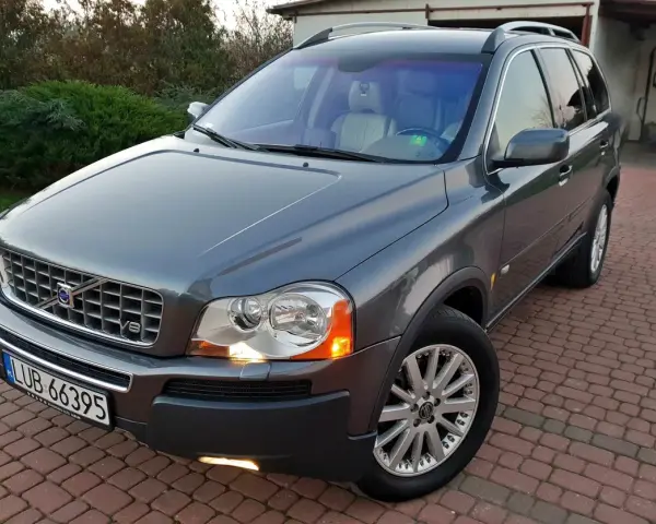 Szary Volvo XC90 z silnikiem V8, zaparkowany na podjeździe z kostki brukowej, otoczony zielenią.