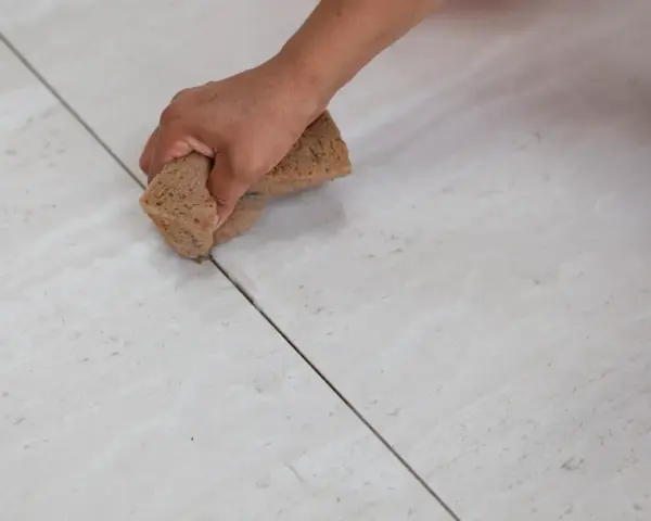 Mano che stuccare piastrelle con una spugna, rifinendo le fughe di un pavimento chiaro.