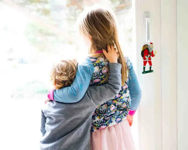 Zwei Kinder umarmen sich vor einem Fenster. Das jüngere Kind, das Angst hat, wird vom älteren Kind getröstet. Ein Weihnachtsmann-Anhänger hängt an der Tür.
