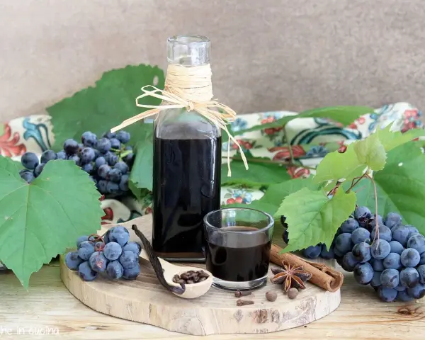 Mosto cotto ricetta: sciroppo scuro in bottiglia e bicchierino, con uva, spezie e cucchiaio di semi.