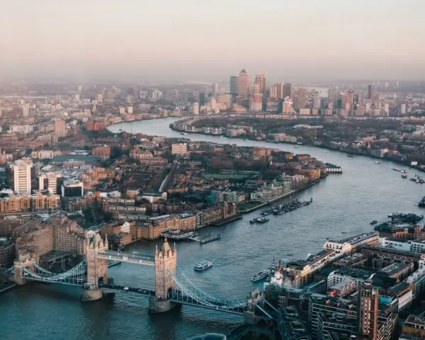 Stunning drone photography and videography captures London's iconic Tower Bridge and the winding Thames River, with the city skyline stretching into the hazy distance.