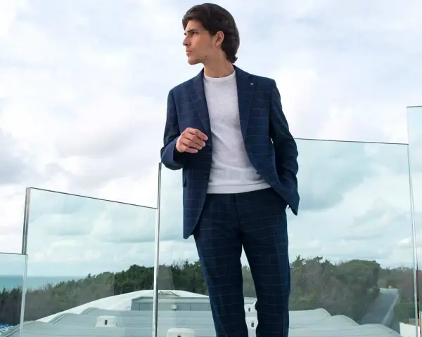 Uomo con giacca blu a quadri, pantalone grigio e scarpe nere, guarda verso l'orizzonte da una terrazza con vista mare.