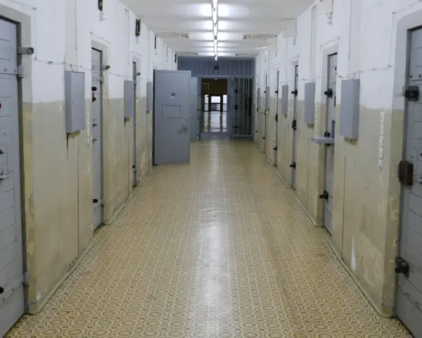 Korytarz więzienny z celami, gdzie może odbywać się tymczasowe aresztowanie. Widać drzwi z numerami, kraty i metalowe zabezpieczenia.