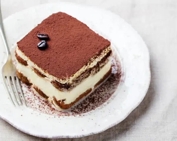 Una fetta di tiramisù, preparata con 500 g di mascarpone, guarnita con cacao e chicchi di caffè, su un piatto bianco con forchetta.