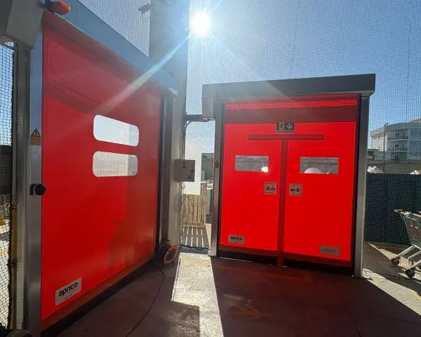 Due porte rosse automatiche, una aperta, mostrano come alzare una porta dal pavimento. Luce solare intensa.