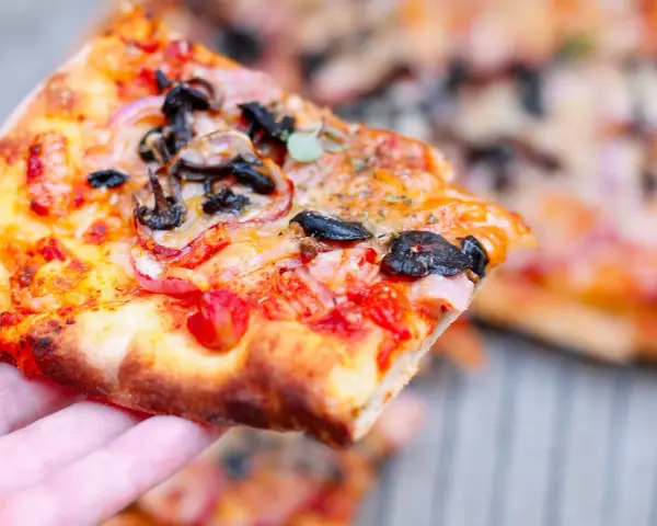 Kawałek pysznej pizzy funghi z pieczarkami, cebulą i pomidorami, trzymany w dłoni.