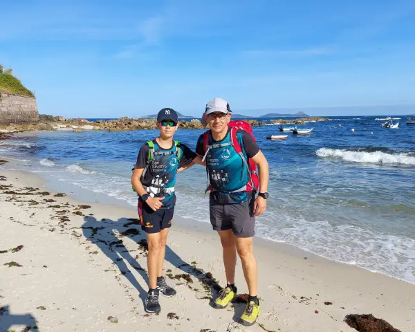 Dos excursionistas, padre e hijo, disfrutan de la playa en la etapa Baiona Vigo. El mar azul y el cielo despejado acompañan su aventura.